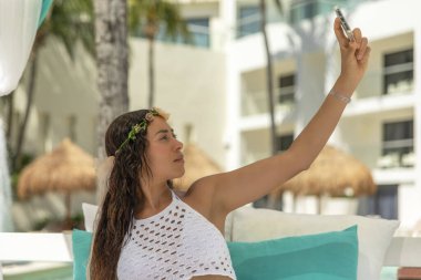 Sexy young female tourist enjoying a relaxing vacation lifestyle at a Caribbean resort, while taking a selfie with her mobile phone. clipart