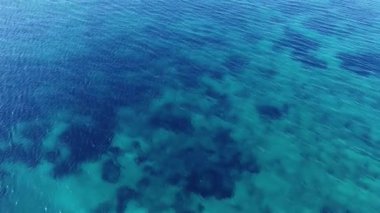 Video yukarıdan denizi sakin ve net bir şekilde gösteriyor. Su derin mavi ve turkuazdır, görünür desenleri ve gölgeleri sualtı manzarası ve mercan oluşumları tarafından oluşturulmuştur..