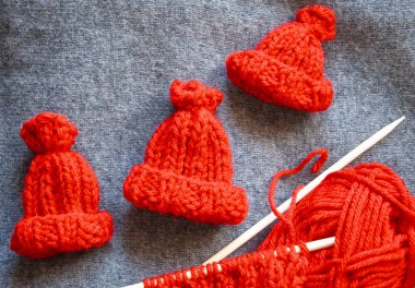 Three small red elf hats, perfect for gnomes, elves, or Santa's helpers, displayed alongside an unfinished knitting project. The scene evokes a festive, whimsical holiday atmosphere, ideal for Christmas crafts, seasonal DIY projects. clipart