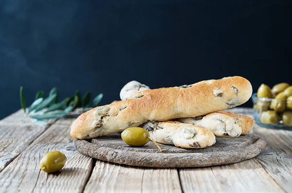 Italian bread sticks with green olives on cutting board, olive tree branch, olives in bowl on wooden table with dark background. Rustically concept of mediterranean cuisine traditions. Copy space