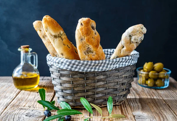 Italian bread sticks with green olives in basket, olive oil, olive tree branch, olives in bowl on wooden table with dark background. Rustically concept of Italian cuisine traditions