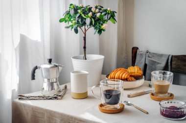 Spring cozy breakfast concept. Two mugs with coffee and milk, croissants, plant with flowers, creamer, coffee pot, jam. Morning light through curtains, linen tablecloth. Kinfolk style