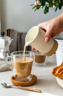 Spring cozy breakfast concept. Two glasses with coffee and milk, croissants, coffee pot, woman hand is holding a creamer and pouring cream to the mug.Vertical banner