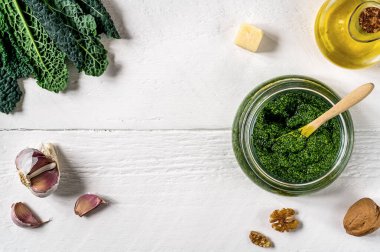 Beyaz ahşap yüzeyde, içinde lahana yaprağı, ceviz, parmesan, sarımsak ve zeytin yağı bulunan cam bir kasede servis edilen ev yapımı lahana pesto sosu. Metin için alanı kopyala