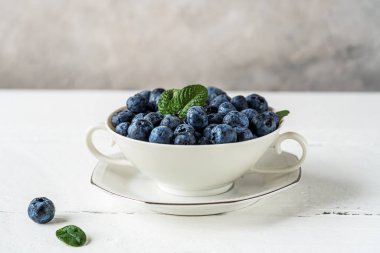 Beyaz ahşap masada beyaz bir çay fincanında taze yaban mersini, gri beton arka plan, yakın plan, yazı için boş alan. Sağlıklı atıştırma konsepti