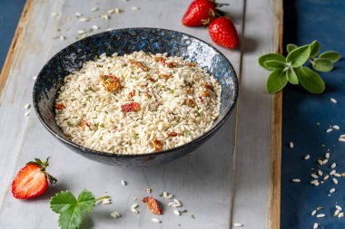 Siyah bir kasede çilek ve otlarla karışık risotto, taze çilek, gri ahşap ve mavi arka planda adaçayı.