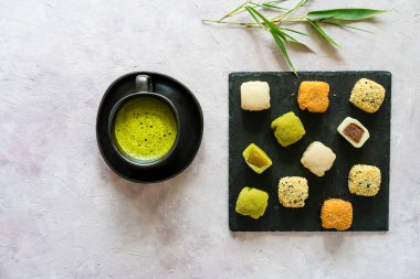 Bir fincan köpüklü kibrit çayı ile kayrak üzerinde çeşitli mochi huzurlu bir düzenleme, bambu yaprakları ile soluk bir arkaplan üzerine kurulmuş