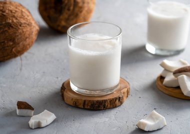 Ahşap altlıklarda iki bardak taze hindistan cevizi sütü ve dokulu bir yüzeye konmuş hindistan cevizi parçaları..