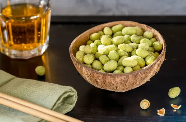 Siyah bir yüzey üzerinde wasabi kaplı fıstıklarla dolu hindistan cevizi kasesi, yanında yeşil bir peçete, yemek çubukları ve bir bardak altın içecekle canlı ve şık bir aperatif oluşturur.