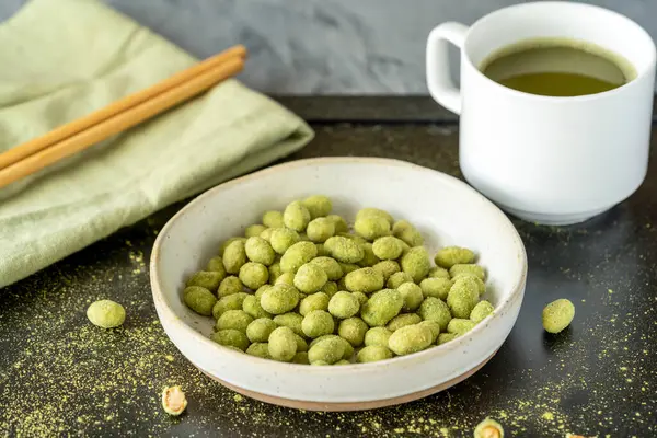 Bir fincan yeşil çay, yemek çubukları ve yeşil peçeteyle kaplanmış seramik bir kase dolusu wasabi kaplı fıstık, Asya 'dan esinlenilmiş hoş bir yemek sahnesi yaratıyor.