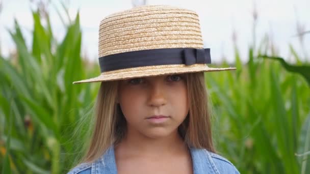 在有机农场玉米地模糊的背景下 一个头戴草帽 严肃的小女孩在镜头前的画像 小女孩站在草地上 把她长长的金发直了过来 — 图库视频影像