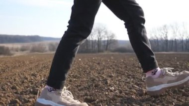 Female feet of small kid goes on the dry ground along ploughed meadow at sunny day. Legs of little girl in sneakers walks on the dirty soil over empty plowed plantation at early spring. Low view.