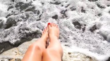 Deniz kıyısındaki taşın üzerinde oturan ince bacaklı kadını kapatın. Yaz tatili boyunca okyanus kıyısında dinlenen tanınmayan bir kız. Güneşli günlerin tadını çıkaran bir kadın ve sıcak deniz dalgaları taşlarda kırılıyor..