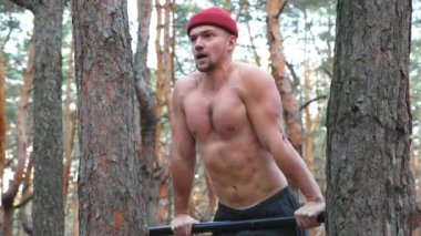 Güçlü ve kaslı bir adam ormanda barfiks çekiyor. Doğada spor yapan cesur bir sporcu. Açık havada duygusal yüz eğitimi almış atletik bir adam. Spor ve aktif yaşam tarzı kavramı.