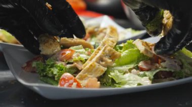 Erkek kolları mutfakta taze malzemelere kızarmış tavuk parçaları koyuyor. Eldivenli şefin elleri Sezar salatasını servis için düzenliyor. Yemek pişirme kavramı. Yavaşla..