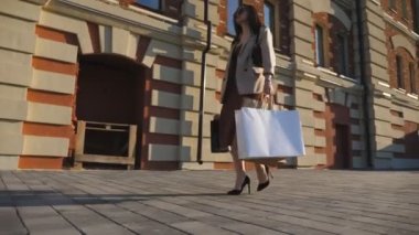 Modaya uygun bayan müşteriler şehir caddesinde kağıt paketler taşıyorlar. Çekici bir stil kızı, alış veriş sonrası kaldırımda yürürken alışveriş torbaları tutuyor. Alışverişkolik kavramı. Yavaşça kapat.