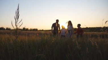 Anne ve babası ve küçük çocukları gün batımında çim tarlalarında el ele yürüyorlar. İki çocuklu mutlu bir aile yaz otlaklarında geziyor ve birlikte vakit geçiriyorlar..