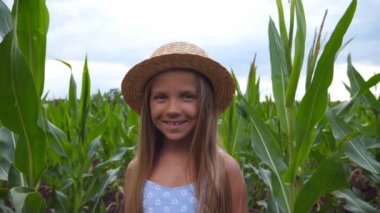 Beautiful little smiling girl in straw hat looking into camera against the background of corn field at organic farm. Portrait of happy small kid with long blonde hair standing in the meadow. Close up.