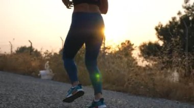 Arka planda güneş doğarken kır yolunda koşan aktif genç bir kızın ayakları. Dişi bacaklar dışarıda koşuyor. Sabahın erken saatlerinde spor yapan bir kadın. Sağlıklı yaşam tarzı kavramı. Yavaş çekim.