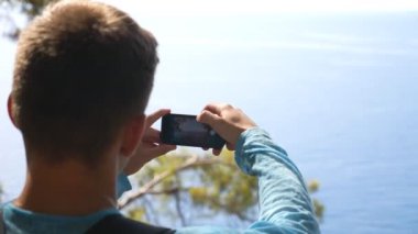 Tanımlanamayan bir adam akıllı telefonuyla deniz manzaralı bir manzaranın fotoğrafını çekiyor. Tepeden güzel deniz resimleri çekmek için telefonu kullanan genç bir adam. Erkek yürüyüşçü yaz tatilinde seyahat etmekten hoşlanır. Yavaş çekim.