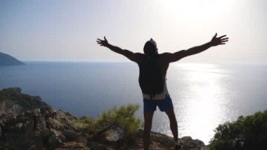 Erkek yürüyüşçü ellerini kaldırarak dağda duruyor ve deniz manzarasına hayran kalıyor. Sırt çantalı bir adam yolculuk sırasında aktif olarak dinleniyor ve tepenin üstünde özgürlüğün tadını çıkarıyor. Arka planda güzel bir manzara. Yavaş çekim