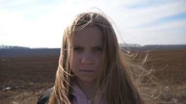 Küçük bir bayan çocuğun portresi organik çiftlikteki tarlanın arka planında kameraya bakıyor. Küçük ciddi kız tarlada duruyor ve uzun sarı saçları rüzgarda savruluyor. Kapat..