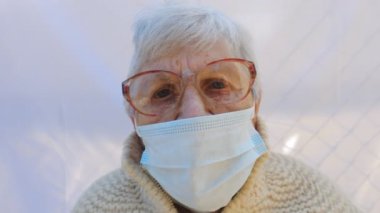 Virüsten koruyucu maske takan büyükannenin portresi. Hüzünlü duygular gösteren yaşlı bir kadın kameraya bakıyor. Salgından korunan sağlık ve güvenlik kavramı. Yaşlılar için koronavirüs karantinası.