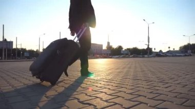 Günbatımında şehir sokağında bavuluyla yürüyen siyah takım elbiseli başarılı iş adamının profili. Kendine güvenen genç adam havaalanı terminaline gidiyor ve tekerlekli bavulu çekiyor. Yan Görünümü Kapat.