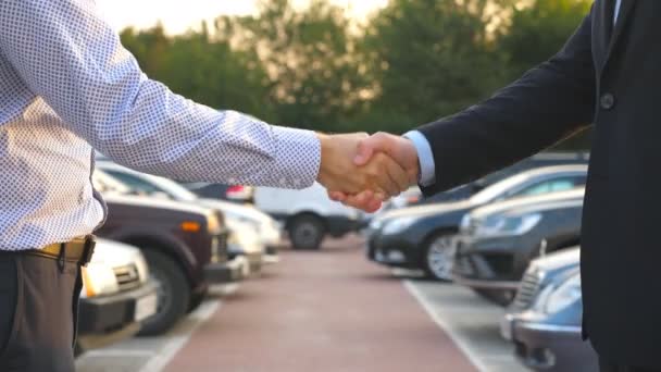 2人のビジネスマンが背景に駐車場で車で互いに握手 マネージャーまたはディーラーとクライアントの間の握手 外で男の腕を振る スローモーションを閉じます — ストック動画