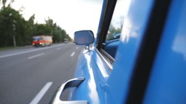 Hareket halindeki eski arabanın sol tarafındaki aynada ciddi bir erkek sürücü yansıması var. Asfalt yolda mavi bir araba kullanan genç bir adam. Arkaplanda odaklanma sorunu var. Arka arkaplan Yavaşça kapat.