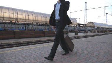 Tanınmayan bir işadamı bavuluyla tren istasyonunda yürüyor ve telefonla konuşuyor. Genç iş adamı demiryolu kenarında geziniyor ve iş görüşmesi yapıyor. Seyahat konsepti. Yavaş çekim.