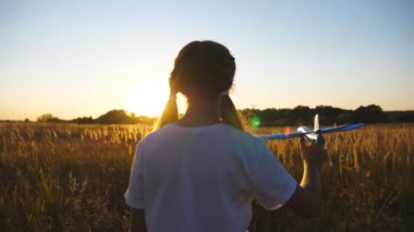 Küçük kız çayırların arasında uçakla oynuyor. Tasasız küçük çocuk arka planda gün batımına karşı oyuncak uçakla sahada yürüyor. Hafta sonunun tadını çıkaran tatlı bir bayan çocuk. Arka görünüm.