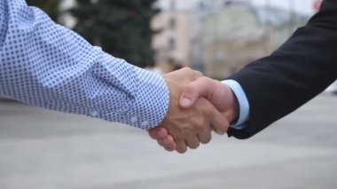 Şehir ortamında birbirini selamlayan iki iş adamı. Bulanık şehir geçmişine sahip genç erkek arkadaşlar el sıkışıyor. İş ortaklarının el sıkışması. Yan Görünüm Yavaşça Kapat.