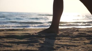 Sahilde altın kumda yürürken arka planda okyanus dalgaları olan kadın ayaklarına yakın dur. Genç kadın bacakları kuma basıyor. Deniz kıyısındaki çıplak ayaklı kız. Yaz tatili. Yavaş çekim.