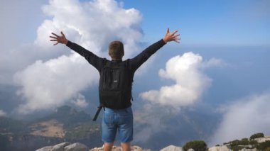 Boy tourist with backpack standing on the edge of the hill and victoriously outstretching hands. Cloudy sky at background. Rear back view Slow motion.