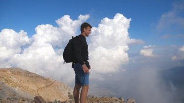Young male hiker with backpack standing on mountain peak and enjoying beautiful landscape. Boy tourist victoriously raising hand on the hill with cloudy sky at background. Travel concept. Slow motion.