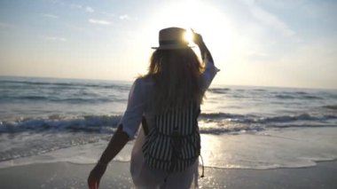 Young woman in shirt with backpack jogging to the sea and raising hands. Beautiful girl in hat running to the shoreline and spraying water with her feet. Happy female enjoying freedom. Slow motion.