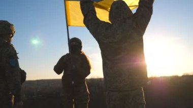 Soldiers of ukrainian army standing at top of hill and lifted up flag of Ukraine. People in military uniform holding waving flag as symbol of victory against russian aggression. End of war in Europe.