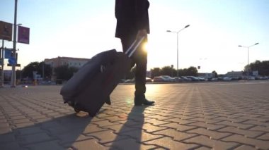 Crane shot of unrecognizable man walking on city street and pulling suitcase on wheels at sunset. Young businessman looking at his watch and going with luggage to airport terminal. Close up Rear view.