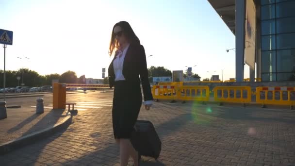 若い女性は日没時に荷物を持って空港駐車場の近くを歩いている ビジネスの女性は街の通りに沿って彼女のスーツケースと行く 背景に太陽のフレア 旅行のコンセプト スローモーション閉じる — ストック動画