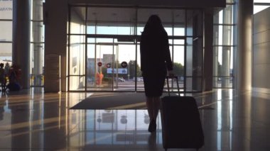 Topuklu giyen genç bir kadın bavuluyla havaalanı koridorunda ya da bekleme odasında yürüyor. Terminalden bagajıyla çıkan iş kadınını takip edin. Seyahat ya da tatil seyahati konsepti. Yavaş çekim.