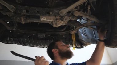 Üniformalı genç bir tamirci atölyedeki bir arabanın süspansiyonunu inceliyor. Garajda kaldırma aracının altında çalışan profesyonel sakallı oto tamircisi. Otomobil hizmeti kavramı. Yavaş çekim.