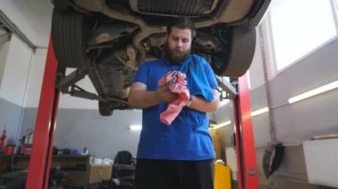 Garajdaki yakıt yağından ellerini silen genç bir tamirci. Üniformalı profesyonel sakallı tamirci atölyede sıkı çalışmasını bitirdi. Araba servisi konseptini düzeltiyorum. Yavaş çekim.