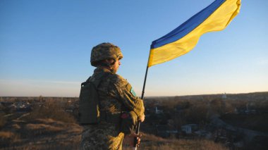 Soldier of ukrainian army stands at hill and holds waving flag of Ukraine. Man in military uniform and helmet lifted up flag against sunset. Victory against russian aggression. Invasion resistance.