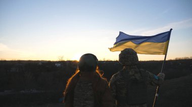 Ukrayna ordusunun askerleri günbatımının arka planına karşı Ukrayna bayrağı çekiyorlar. Askeri üniformalı insanlar sarı-mavi bayrak kaldırdı. Rus saldırısına karşı zafer. İstila direnişi kavramı.