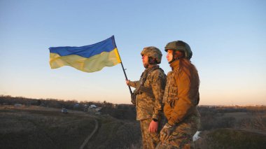Soldiers of ukrainian army standing at the hill and waving flag of Ukraine. People in military uniform and helmet lifted up yellow-blue flag. Victory against russian aggression. Invasion resistance.