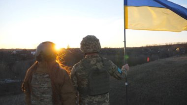 Ukrayna ordusunun askerleri günbatımının arka planına karşı Ukrayna bayrağı çekiyorlar. Askeri üniformalı insanlar sarı-mavi bayrak kaldırdı. Rus saldırısına karşı zafer. İstila direnişi kavramı.