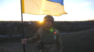 Ukrayna bayrağı sallayan Ukrayna ordusunun askeri. Askeri üniformalı ve kasklı bir adam gün batımında bayrak kaldırdı. Rus saldırısına karşı zafer. İstila direnci konsepti. Yavaş çekim.