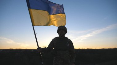 Ukrayna bayrağı sallayan Ukrayna ordusunun askeri. Askeri üniformalı ve kasklı bir adam gün batımında bayrak kaldırdı. Rus saldırısına karşı zafer. İstila direnci konsepti. Yavaş çekim.