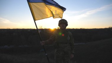 Ukrayna bayrağı sallayan Ukrayna ordusunun askeri. Askeri üniformalı ve kasklı bir adam gün batımında bayrak kaldırdı. Rus saldırısına karşı zafer. İstila direnci konsepti. Yavaş çekim.